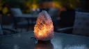 Generate a 1_1 square image of a Grey Himalayan Salt Lamp with natural irregular shape, placed on an outdoor patio table during evening time. Use soft ambient outdoor lighting and warm glow from the lamp. Backgrou.webp